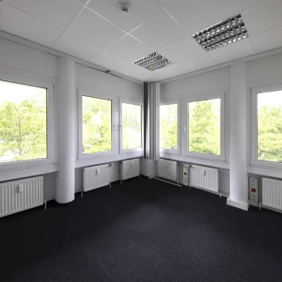 Das Bild zeigt ein modernes Eckbüro mit großen Fensterflächen, Teppichboden und hellem Tageslicht.