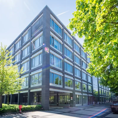Das Bild zeigt ein modernes Bürogebäude an einer Straßenecke in Eschborn, umgeben von Bäumen und Gehwegen.