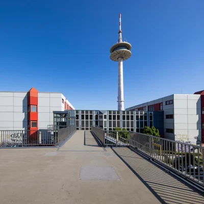 Auf dem Bild ist eine Dachterrasse mit Verbindungsbrücke zwischen zwei Bürogebäuden und einem Fernsehturm im Hintergrund zu sehen.
