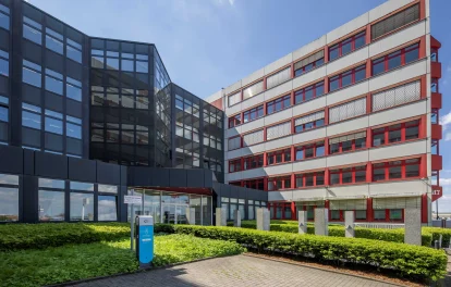 Das Bild zeigt ein modernes Bürogebäude mit Glasfassade und rotem Fensterrahmen in einem Gewerbegebiet.