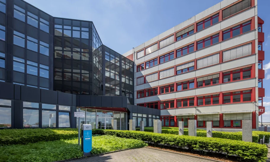 Das Bild zeigt ein modernes Bürogebäude mit Glasfassade und rotem Fensterrahmen in einem Gewerbegebiet.