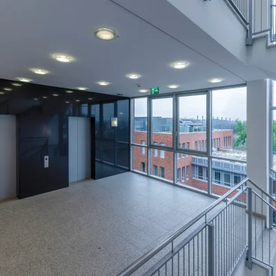 Das Bild zeigt ein modernes Treppenhaus mit Aufzug und großer Glasfront in einem Bürogebäude in Eschborn mit Blick auf andere Gebäudeteile und viel Grün.