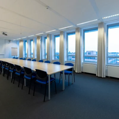 Auf dem Bild ist ein heller Konferenzraum in einem Bürogebäude in Eschborn mit langer Tischreihe, Fensterfront, Beamer und Leinwand zu sehen.