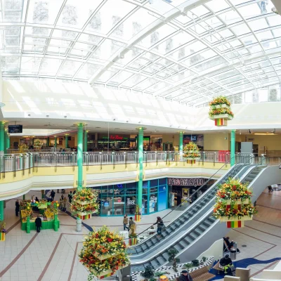 Zu sehen ist das lichtdurchflutete Innere des Einkaufszentrums mit Rolltreppen, Glasdach und floraler Dekoration.