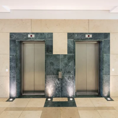 Das Bild zeigt den Aufzugsbereich im Inneren des Next Office Bürogebäudes in München mit Steinverkleidung und zwei Fahrstühlen.