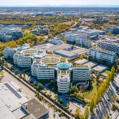 Das Bild zeigt den Bürokomplex Next Office in München aus der Luft, eingebettet in ein Gewerbegebiet mit umliegenden Gebäuden und herbstlicher Vegetation.