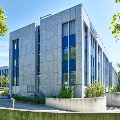 Mehrteiliger Bürokomplex mit blauer Glasfassade, Innenhof und angrenzenden Parkplätzen, umgeben von Bäumen.