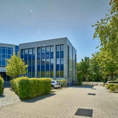 Außenaufnahme eines modernen Bürogebäudes mit blauer Glasfassade, Parkplatzflächen und gepflegten Grünanlagen.