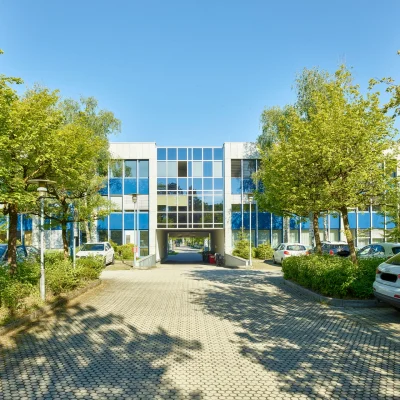 Das Bild zeigt ein Bürogebäude mit blauer Glasfassade, Zufahrt und Parkflächen, umgeben von gepflegten Bäumen und Grünanlagen.