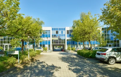 Das Bild zeigt ein Bürogebäude mit blauer Glasfassade, Zufahrt und Parkflächen, umgeben von gepflegten Bäumen und Grünanlagen.