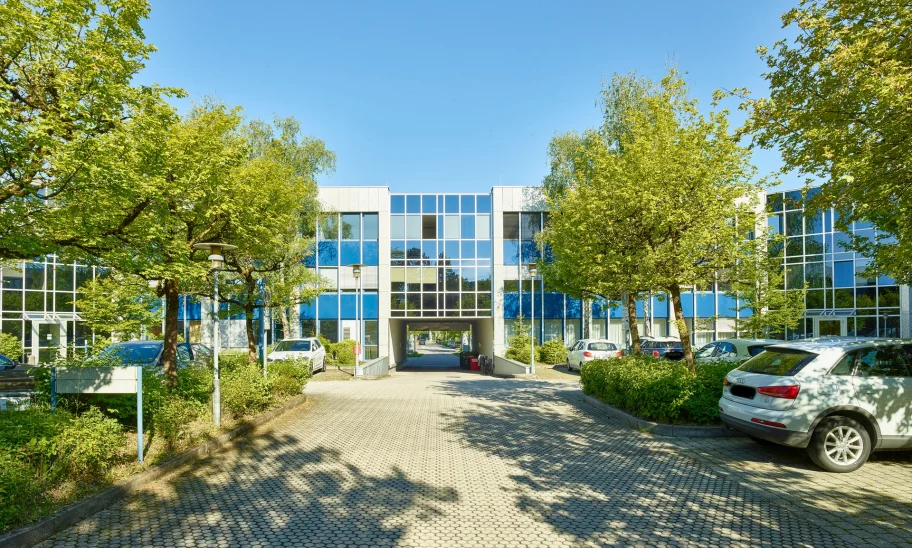Das Bild zeigt ein Bürogebäude mit blauer Glasfassade, Zufahrt und Parkflächen, umgeben von gepflegten Bäumen und Grünanlagen.