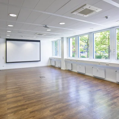 Großzügiger Konferenzraum mit Fensterfront, hellem Holzboden, Deckenleuchten und Projektionsfläche.