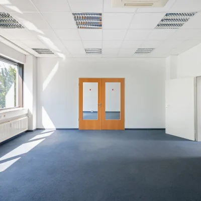 Ein moderner Büroraum mit Schreibtischen, Teppichboden und großer Fensterfläche.