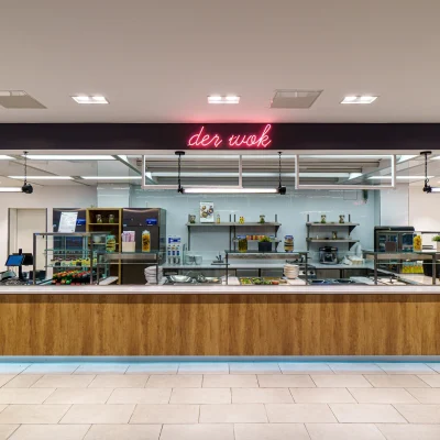 Die Aufnahme zeigt eine moderne Kantine mit einer langen Ausgabetheke, Neonbeschriftung „der wok“ und gut organisierter Küchenzeile.