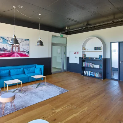 Ein heller Wartebereich mit blauer Couch, Bücherregal, Holzboden und modernen Lampen im industriellen Stil.
