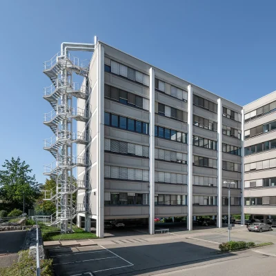 Außenansicht eines modernen Bürogebäudes mit markanter Fassade, Außentreppe und angrenzendem Parkplatz.