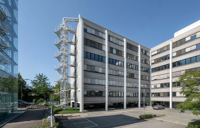 Außenansicht eines modernen Bürogebäudes mit markanter Fassade, Außentreppe und angrenzendem Parkplatz.