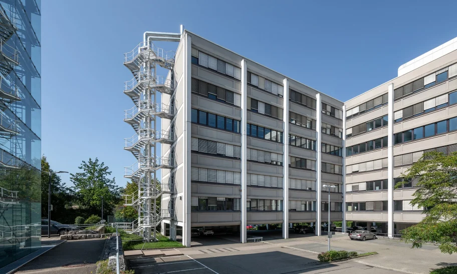 Außenansicht eines modernen Bürogebäudes mit markanter Fassade, Außentreppe und angrenzendem Parkplatz.
