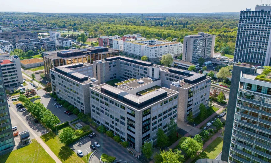 Die Luftaufnahme zeigt ein modernes Bürogebäude im Frankfurter Stadtteil Niederrad mit umliegenden Grünflächen und Nachbargebäuden.