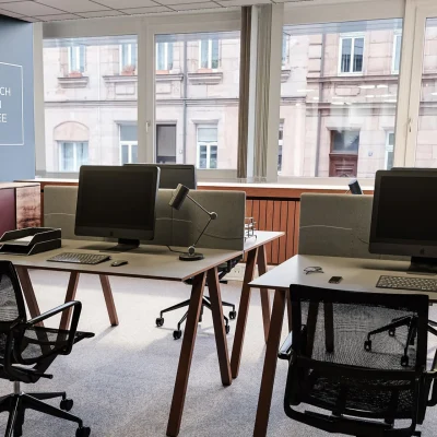 Ein moderner Büroraum mit mehreren Arbeitsplätzen, Computern und Blick auf das gegenüberliegende Gebäude.