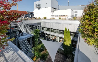 Ein moderner Büro-Innenhof mit Terrasse, Glasfassade, Metalltreppe und umliegender Stadtbebauung.