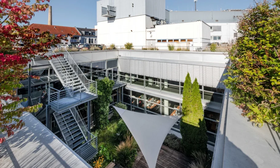Ein moderner Büro-Innenhof mit Terrasse, Glasfassade, Metalltreppe und umliegender Stadtbebauung.