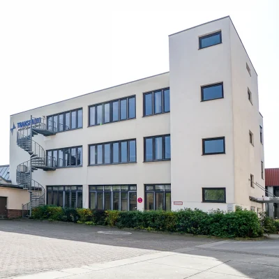 Abgebildet ist ein modernes Bürogebäude mit Wendeltreppe und Stellplätzen vor dem Eingang in einem Gewerbepark.