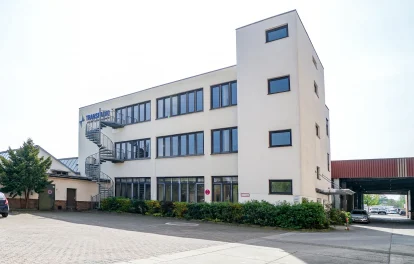 Abgebildet ist ein modernes Bürogebäude mit Wendeltreppe und Stellplätzen vor dem Eingang in einem Gewerbepark.