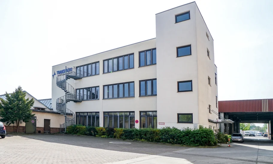 Abgebildet ist ein modernes Bürogebäude mit Wendeltreppe und Stellplätzen vor dem Eingang in einem Gewerbepark.