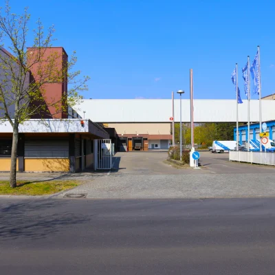Die Aufnahme zeigt den Eingangsbereich eines Gewerbeparks mit Logistikhallen, Zufahrt und Bürotrakt bei sonnigem Wetter.