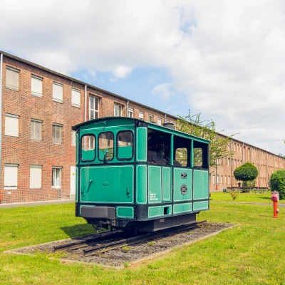 Das Bild zeigt ein Backstein-Bürogebäude im Industriepark Kassel Mittelfeld mit gepflegter Grünfläche, einem Parkplatz und einer kleinen historischen Lokomotive im Vordergrund.