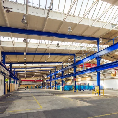Innenansicht einer großen Lagerhalle mit Hochregalen, Lichtkuppeln und blauen Stahlträgern.