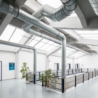 Leerer Innenbereich einer großen Halle mit Oberlichtern und sichtbaren Versorgungsleitungen.