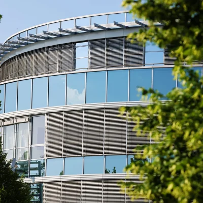 Das Bild zeigt die runde Glasfassade eines modernen Bürogebäudes im Kerpen Techpark, teilweise durch Bäume verdeckt.