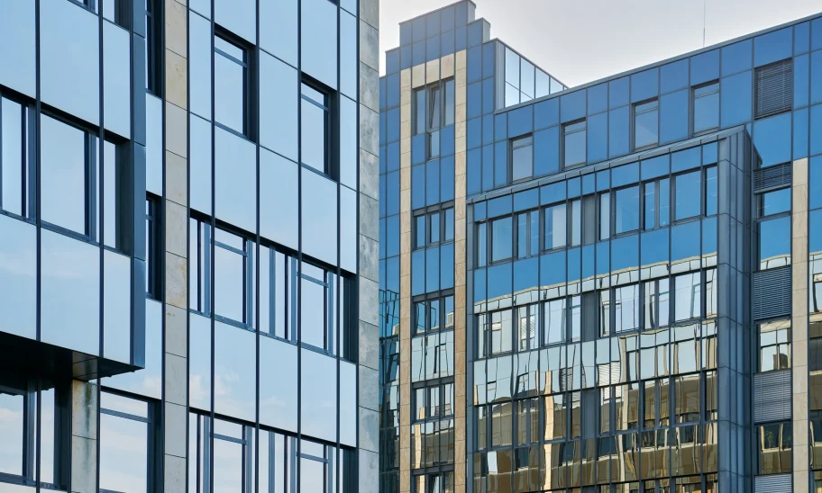 Eine moderne Glasfassade eines Bürohochhauses mit Spiegelung des Himmels und umliegender Gebäude.