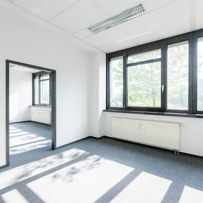 Ein leerer Büroraum mit Durchgang und Fensterfront sowie Teppichböden und Heizungen.