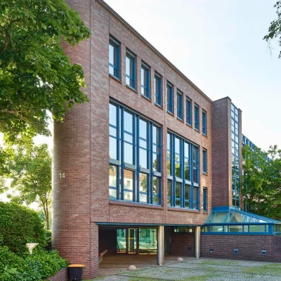 Bürogebäude mit Ziegelfassade und großen Fenstern, umgeben von Bäumen und Grünflächen.Bürogebäude mit Ziegelfassade und großen Fenstern, umgeben von Bäumen und Grünflächen.