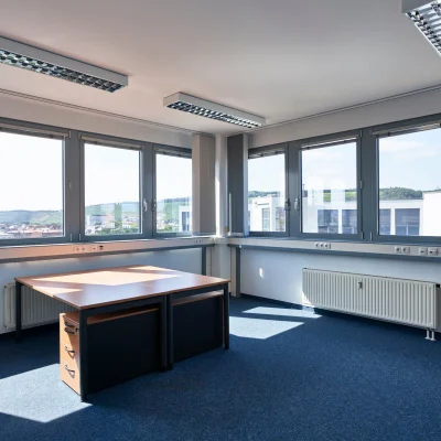 Ein lichtdurchflutetes Großraumbüro mit Teppichboden, mehreren Fenstern und einem Arbeitsplatz mit zwei Schreibtischen.