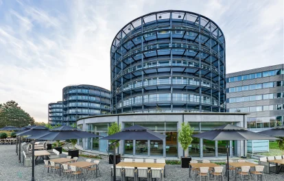 Ein modernes Bürogebäude mit runder Glasfassade in Eschborn und einem großzügigen Außenbereich mit Sitzgruppen und Sonnenschirmen.