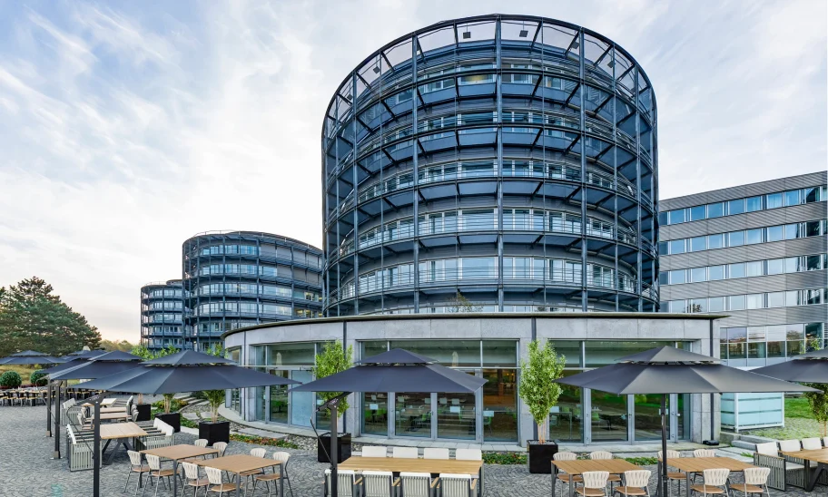 Ein modernes Bürogebäude mit runder Glasfassade in Eschborn und einem großzügigen Außenbereich mit Sitzgruppen und Sonnenschirmen.