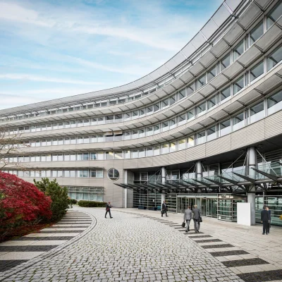 Die Außenansicht eines modernen Bürohauses mit geschwungener Glasfassade und mehreren Personen im Eingangsbereich.