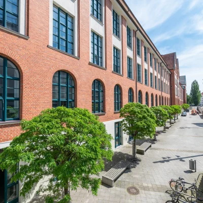 Das Bild zeigt die Außenansicht eines Bürogebäudes mit Backsteinfassade, Fensterreihen und begrüntem Gehweg.