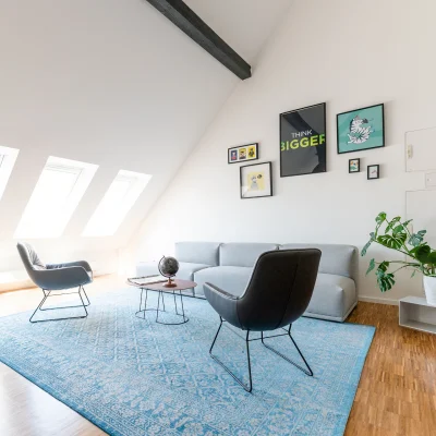Das Bild zeigt eine gemütliche Lounge mit Sofa, Sesseln, Teppich und Pflanzen unter Dachschrägen.