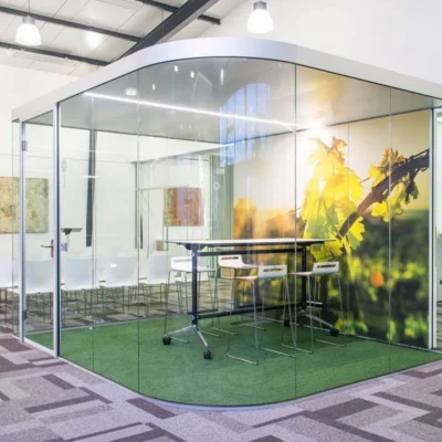 Verglaster Meetingraum mit Naturmotivfolie und hohen Barhockern in einem offenen Bürobereich mit modernem Teppichdesign.