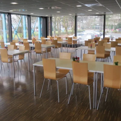 Großzügiger Mitarbeiter- und Aufenthaltsbereich in einem Bürogebäude an der Adickesallee in Frankfurt mit zahlreichen Tischen und Holzstühlen, bodentiefen Fenstern und viel Tageslicht.