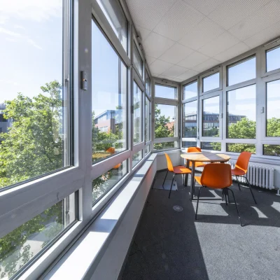 Heller Besprechungsbereich mit großen Panoramafenstern in einem Bürogebäude der Dynamostraße in Mannheim; orange Stühle und ein runder Tisch stehen in einer lichtdurchfluteten Ecke mit Ausblick auf umliegende Bäume und moderne Gebäude.