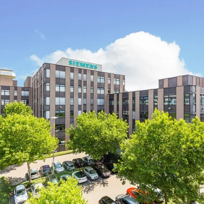 Moderner Bürokomplex in Mannheim mit Siemens-Schriftzug, mehreren sechsgeschossigen Gebäudeteilen und einem großzügigen Parkplatzbereich im Vordergrund, umgeben von grünen Bäumen unter blauem Himmel.