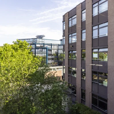 Blick zwischen zwei Bürogebäuden in der Dynamostraße in Mannheim, mit modernen Fassaden aus Glas und Beton sowie dichtem Baumbestand im Vordergrund; sonnige Außenaufnahme mit klarer Sicht auf angrenzende Architektur.
