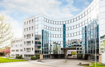 Frontale Außenansicht eines modernen Büroensembles in der Heßbrühlstraße in Stuttgart mit markanter, gebogener Glasfassade, die den Vorplatz sowie umliegende Gebäude widerspiegelt; helle, klare Architektur unter leicht bewölktem Himmel.