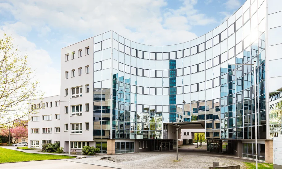 Frontale Außenansicht eines modernen Büroensembles in der Heßbrühlstraße in Stuttgart mit markanter, gebogener Glasfassade, die den Vorplatz sowie umliegende Gebäude widerspiegelt; helle, klare Architektur unter leicht bewölktem Himmel.